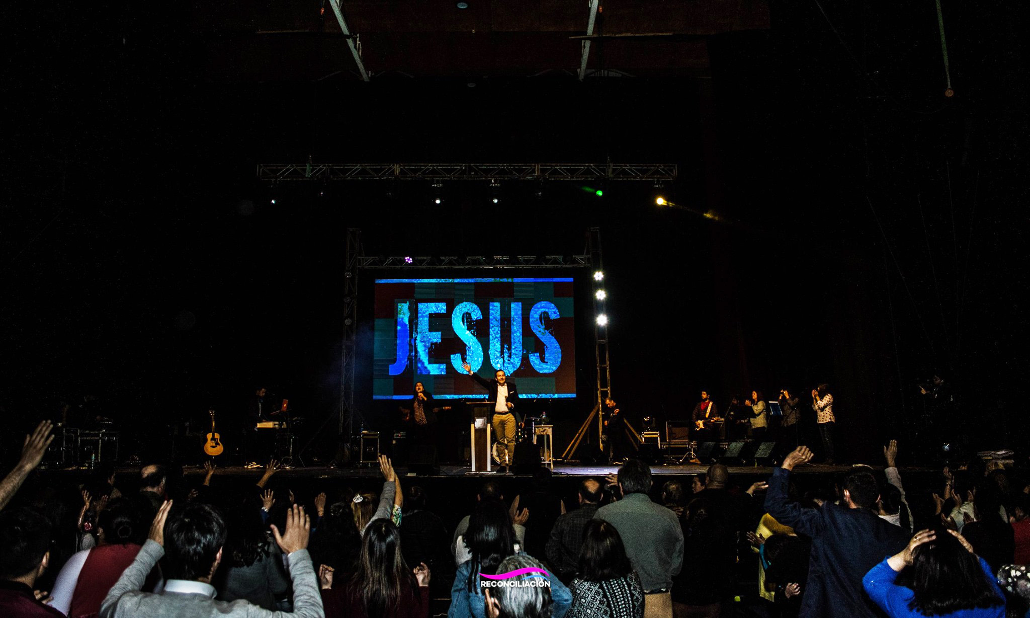 Escenario de una reunión cristiana con la palabra JESUS en pantalla y la congregación adorando.