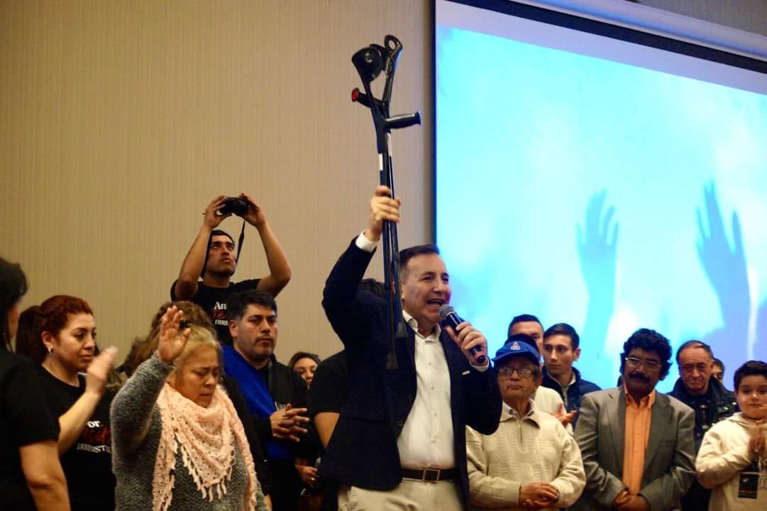 Manuel levantando una muleta durante un testimonio de fe rodeado por la congregación en una reunión cristiana.