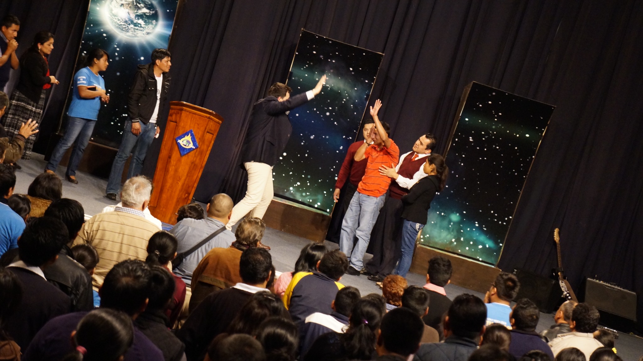Momento de oración en el escenario durante una reunión cristiana con asistentes al fondo.